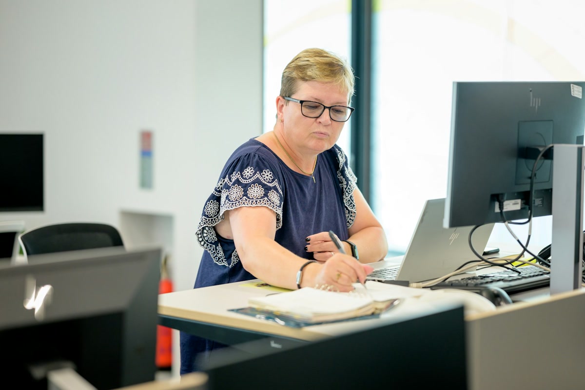 Office worker at a standing desk writing notes beside a laptop